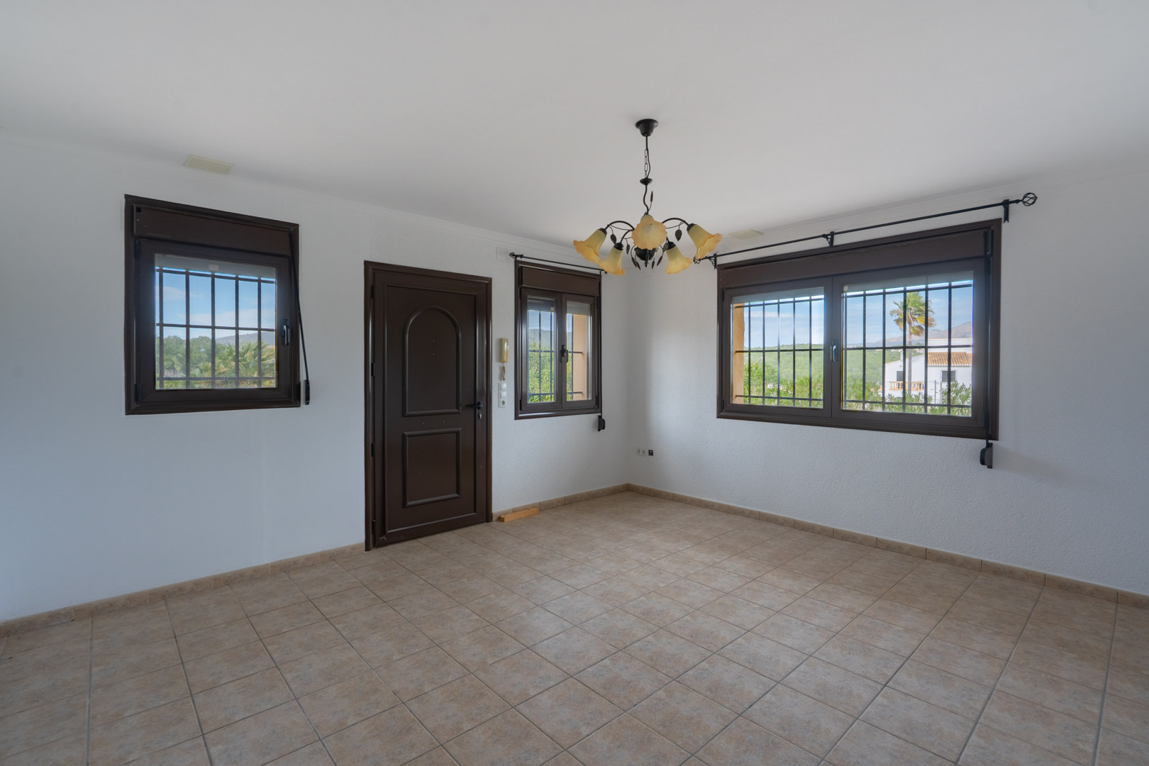 Estancia con suelo cerámico beige, puerta y ventanas oscuras con rejas de seguridad. Incluye lámpara de techo de estilo floral y vistas al paisaje exterior.