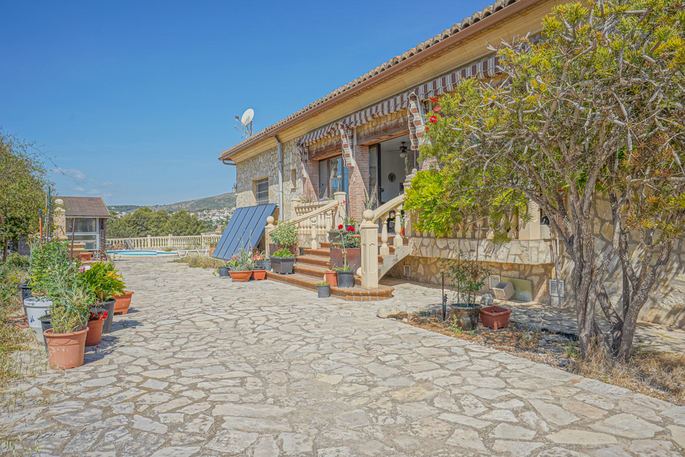 Exterior con patio de piedra natural, fachada de mampostería y paneles solares. Escaleras con balaustrada dan acceso a un porche elevado con toldos.