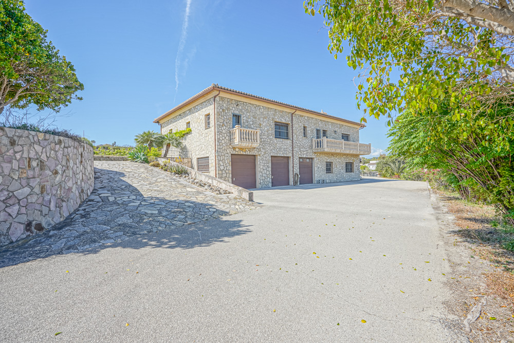 Fachada de piedra natural con tres puertas de garaje, rampa de acceso empedrada y balcones con balaustradas bajo un cielo despejado.