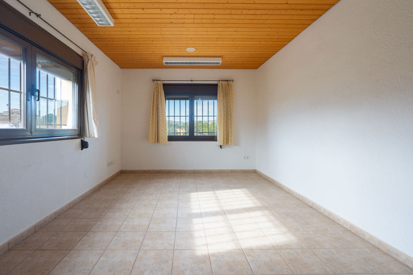 Habitación diáfana con techo de madera natural, suelo de gres beige y ventanas con rejas de seguridad que aportan abundante luz natural.