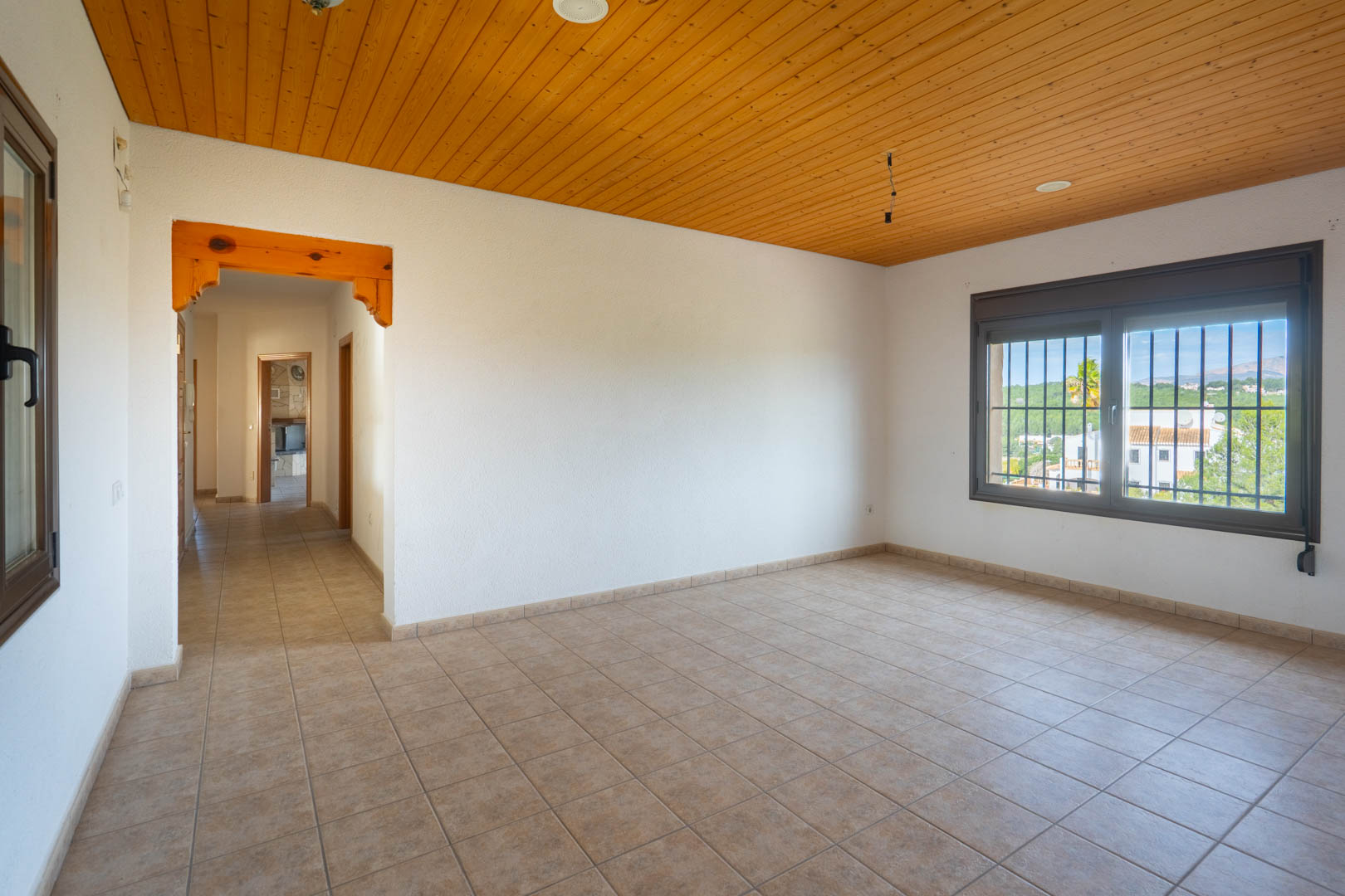 Habitación diáfana con techo de paneles de madera, suelos cerámicos y ventana con rejas que ofrece vistas despejadas al entorno rural.