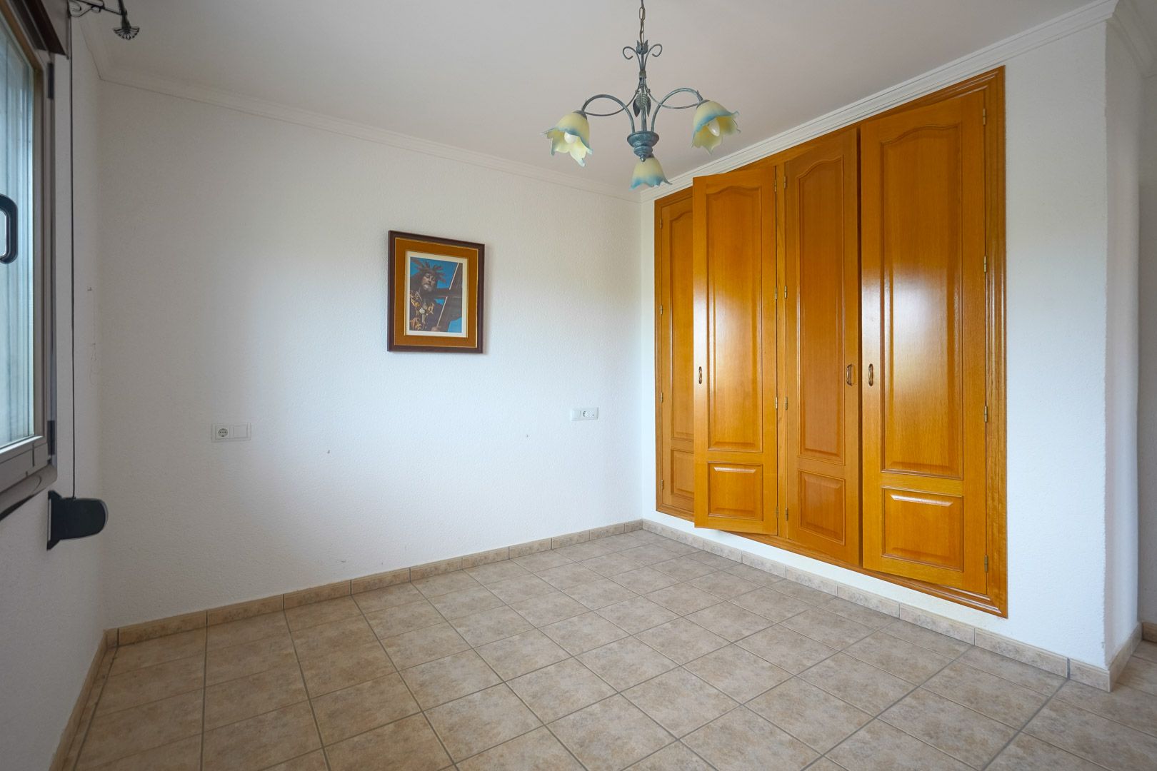 Habitación vacía con armario empotrado de madera de cuatro puertas, suelo de gres cerámico en tonos beige y una lámpara de techo decorativa de estilo floral.