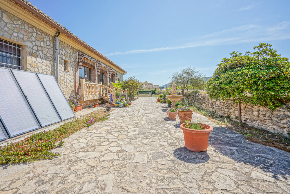 Patio exterior con pavimentación de piedra irregular, fuente ornamental, maceteros de terracota y paneles solares térmicos junto a la fachada de piedra.