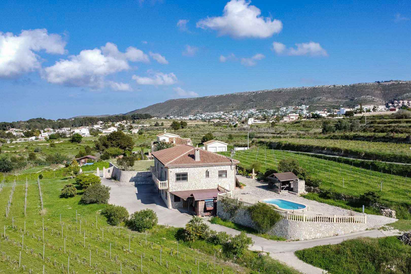 Perspectiva aérea de una villa rústica de piedra con piscina, terraza de piedra con balaustrada y extensos viñedos rodeados de un entorno montañoso.
