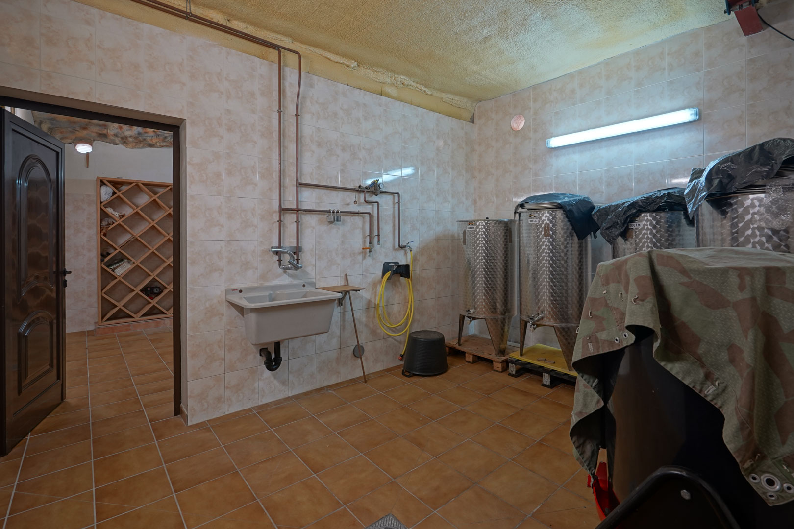 Sala técnica de producción vinícola con depósitos de acero inoxidable, fregadero industrial y tuberías de cobre. Al fondo se aprecia una bodega con estanterías de madera.