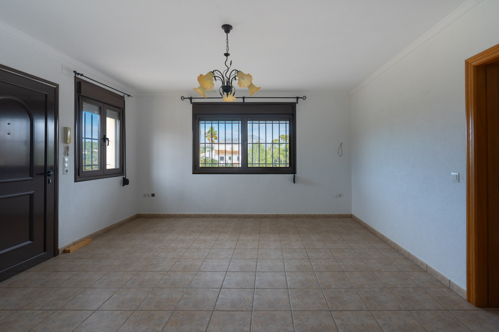 Sala vacía con suelos de gres cerámico, ventanas oscuras con rejas de seguridad y una lámpara de hierro forjado con tulipas de cristal.