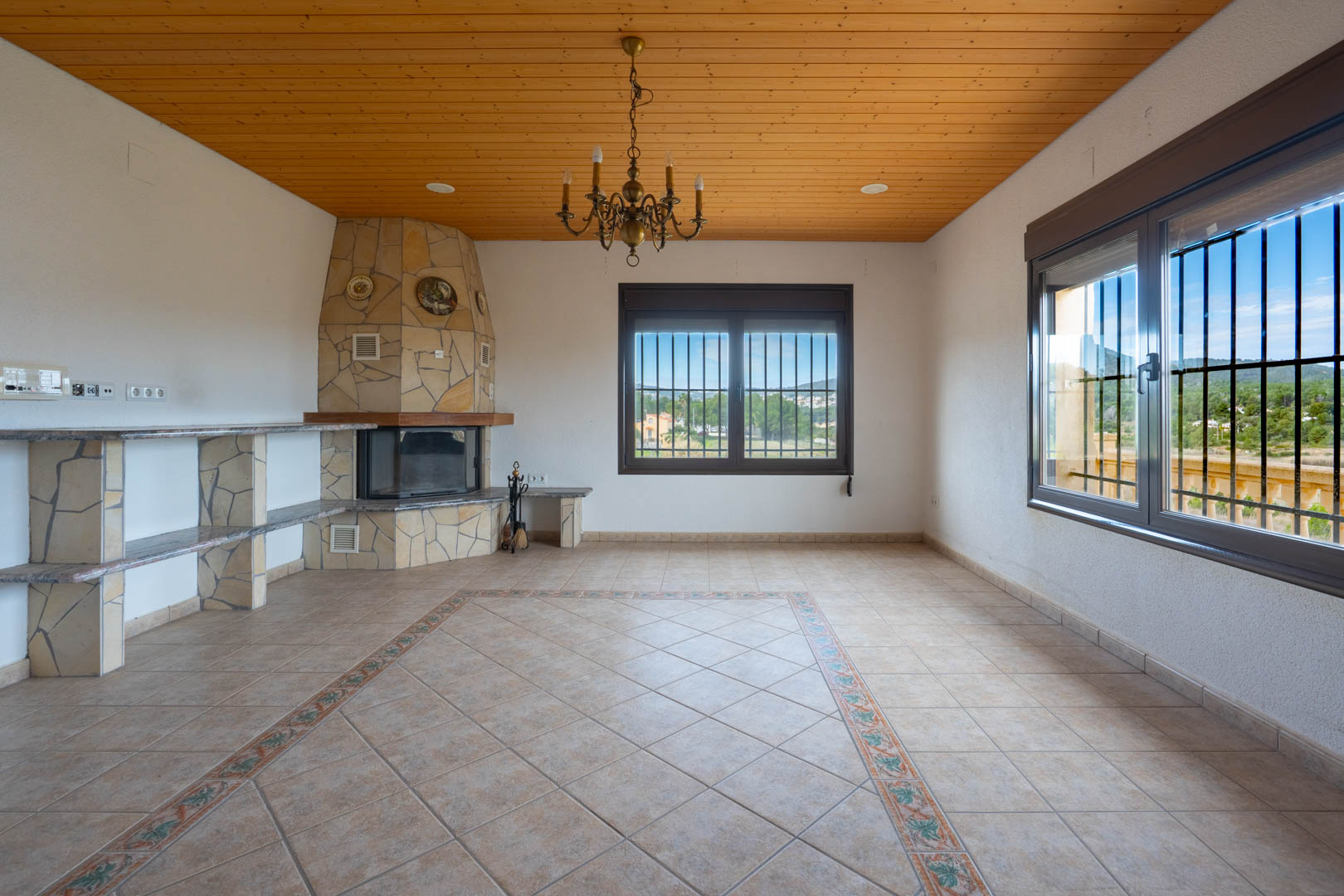 Salón con techo de madera, chimenea revestida en piedra natural y suelos cerámicos con cenefa decorativa. Incluye ventanas con vistas al paisaje.
