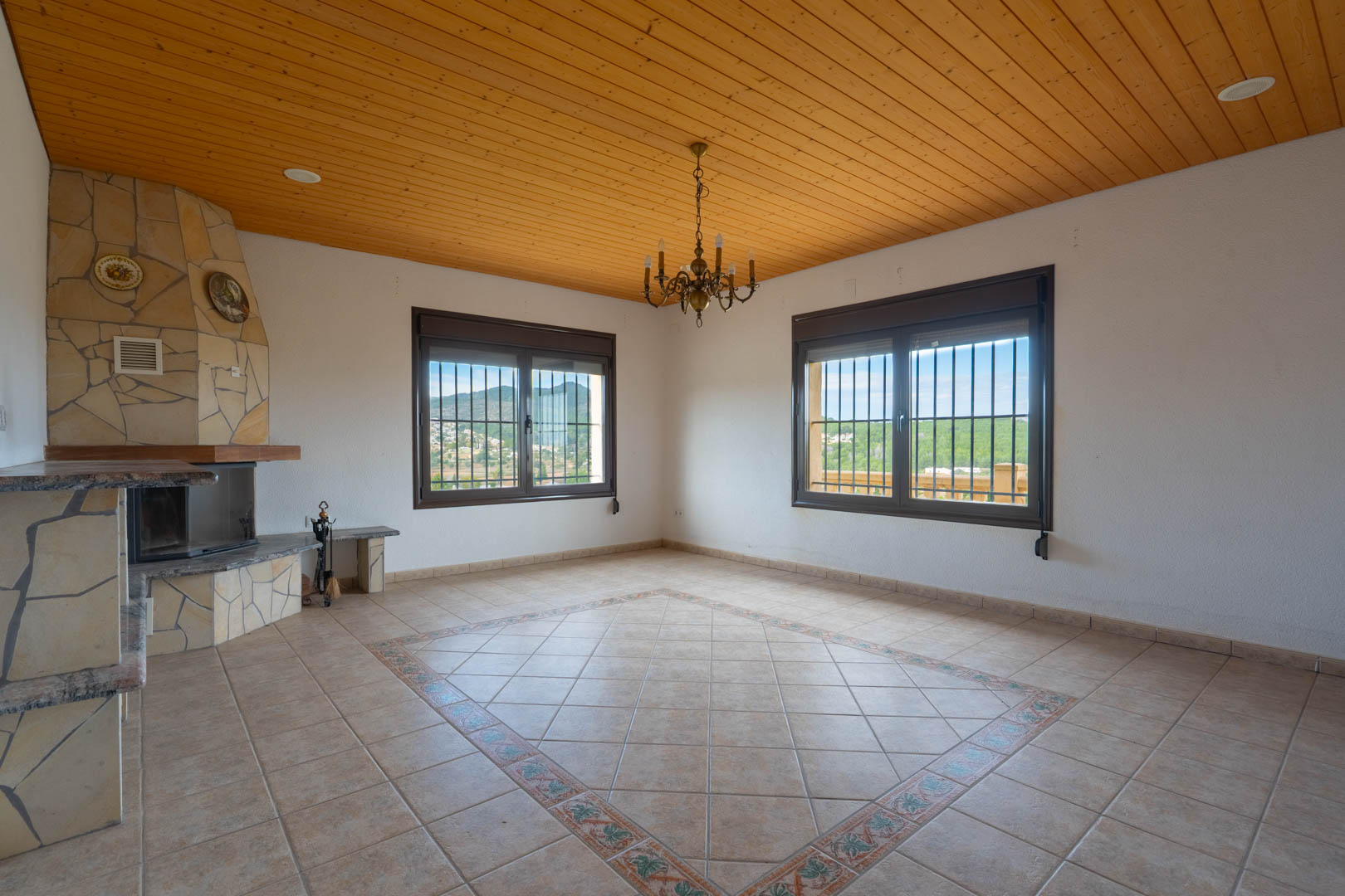 Salón con techo de paneles de madera, chimenea de piedra en esquina y suelo cerámico con cenefa decorativa. Los ventanales ofrecen vistas despejadas a las montañas.