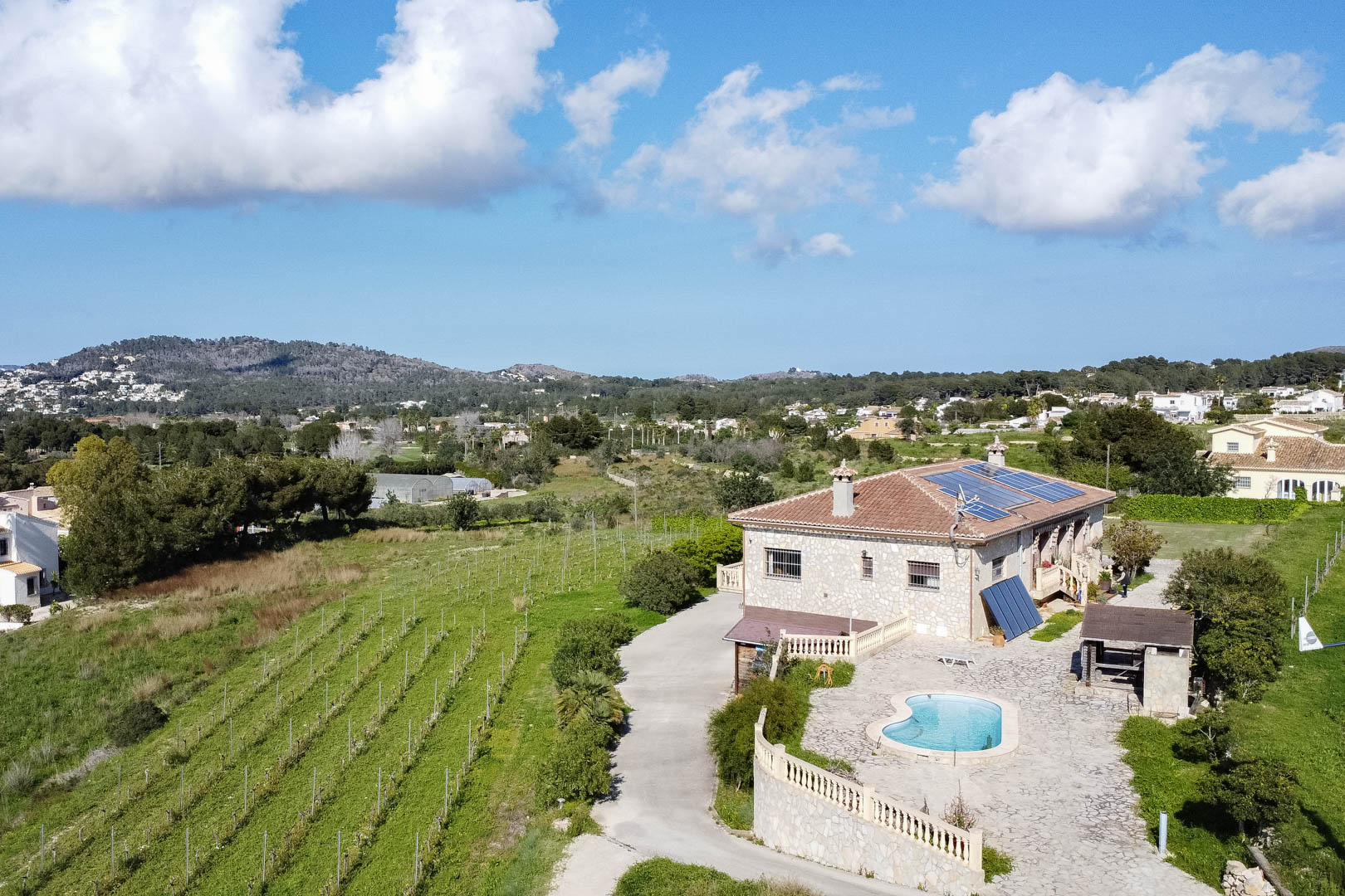 Vista aérea de una casa de campo de piedra con paneles solares, piscina de forma riñón sobre terraza empedrada y viñedos contiguos en un entorno rural.