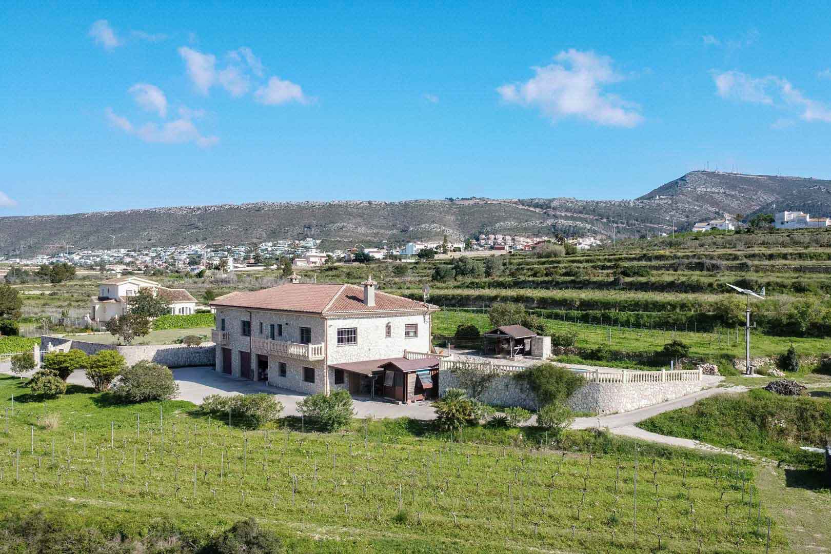 Vista aérea de una finca de piedra con tejado de terracota y balcones, rodeada de viñedos y terrenos verdes bajo un cielo despejado con montañas al fondo.