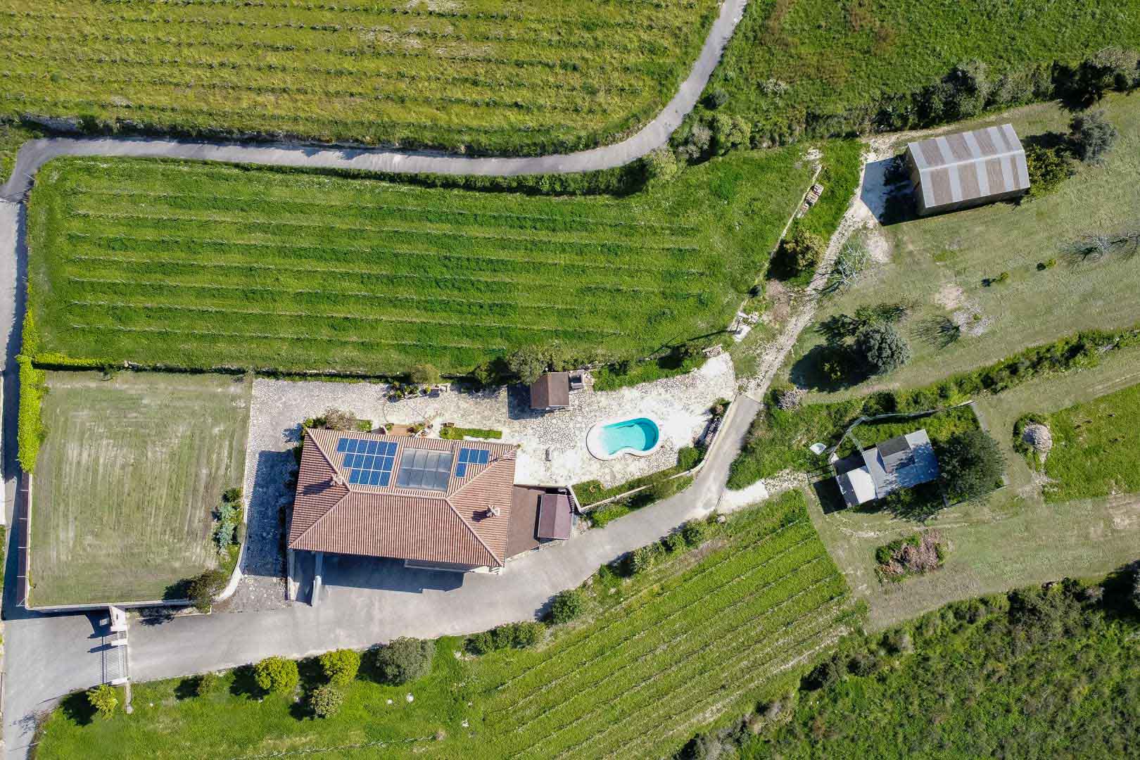 Vista aérea de una propiedad rural con paneles solares en el tejado, una piscina de forma irregular y amplias parcelas agrícolas con cultivos en hileras.