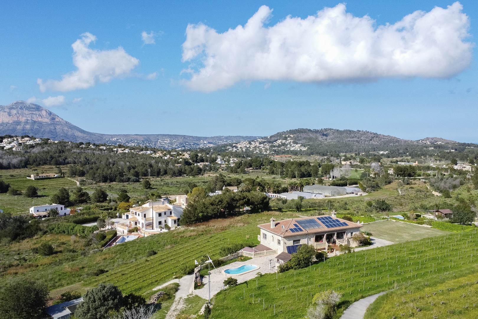 Vista aérea de una villa de piedra con paneles solares, piscina privada y aerogenerador, rodeada de extensos viñedos y un entorno montañoso natural.