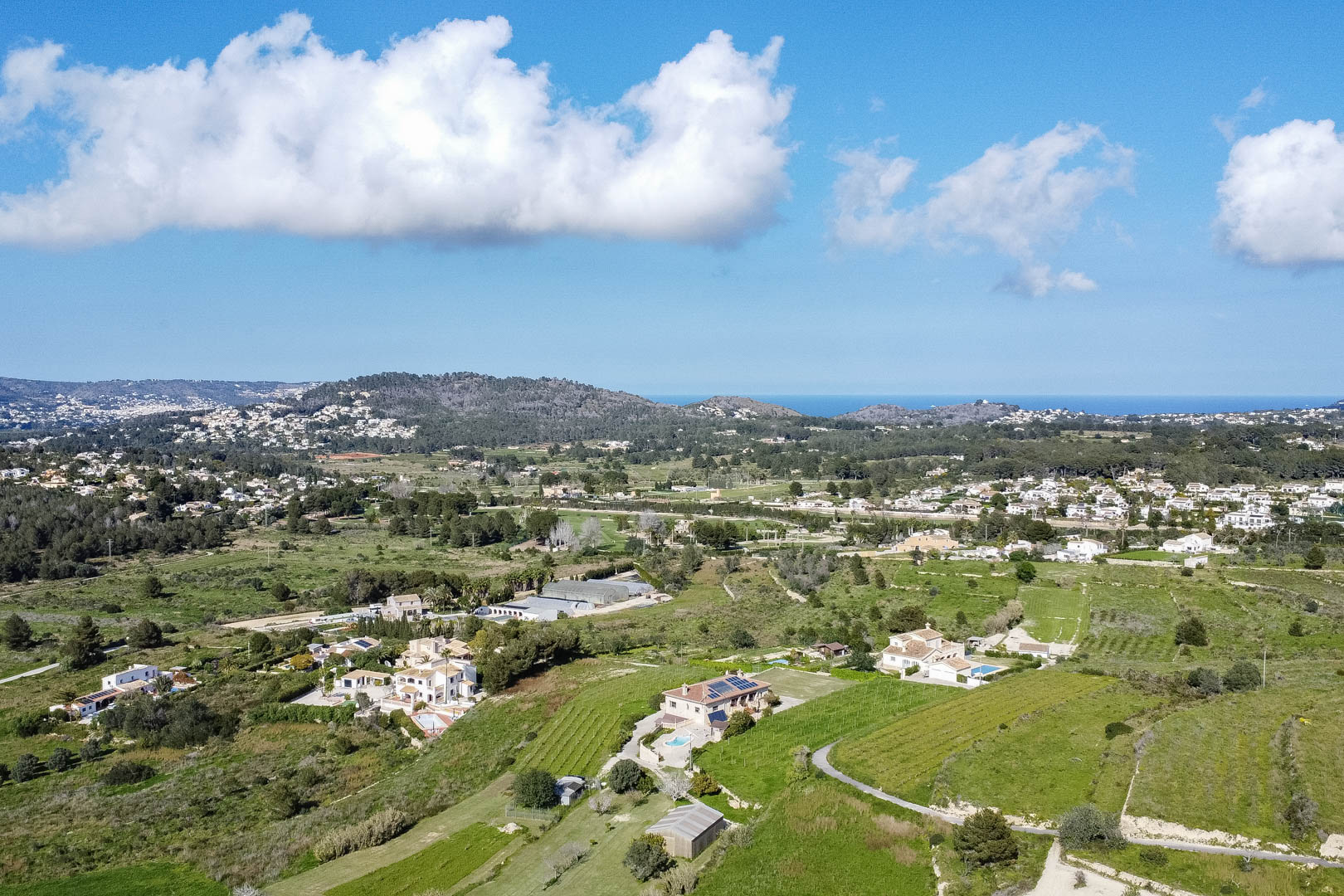 Vista aérea panorámica del paisaje mediterráneo con valles verdes, villas independientes con piscina y vistas lejanas al mar bajo un cielo despejado.