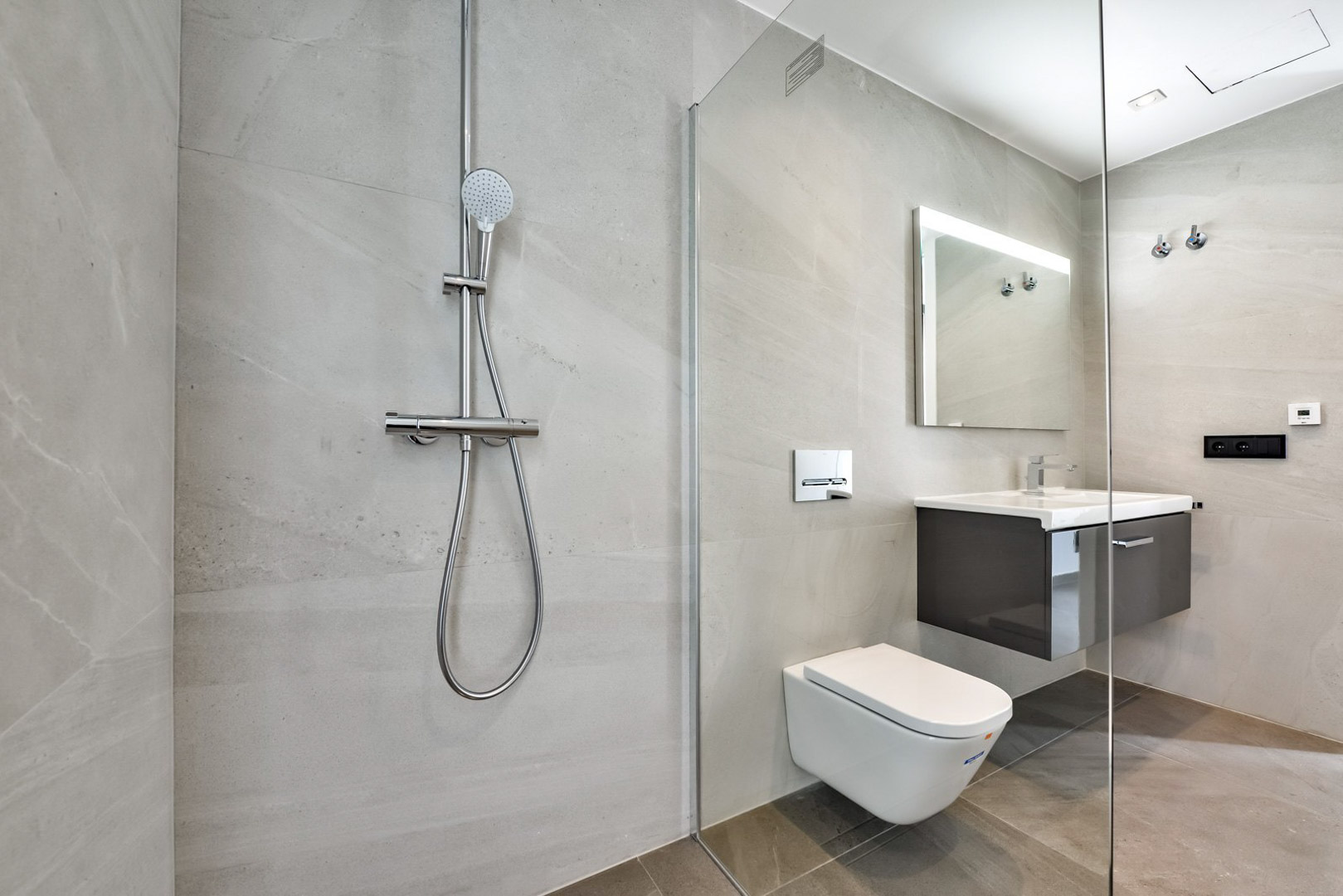 Baño moderno con ducha italiana, mampara de cristal, inodoro suspendido y mueble de lavabo flotante. Revestimiento de gres porcelánico en tonos grises y espejo retroiluminado.