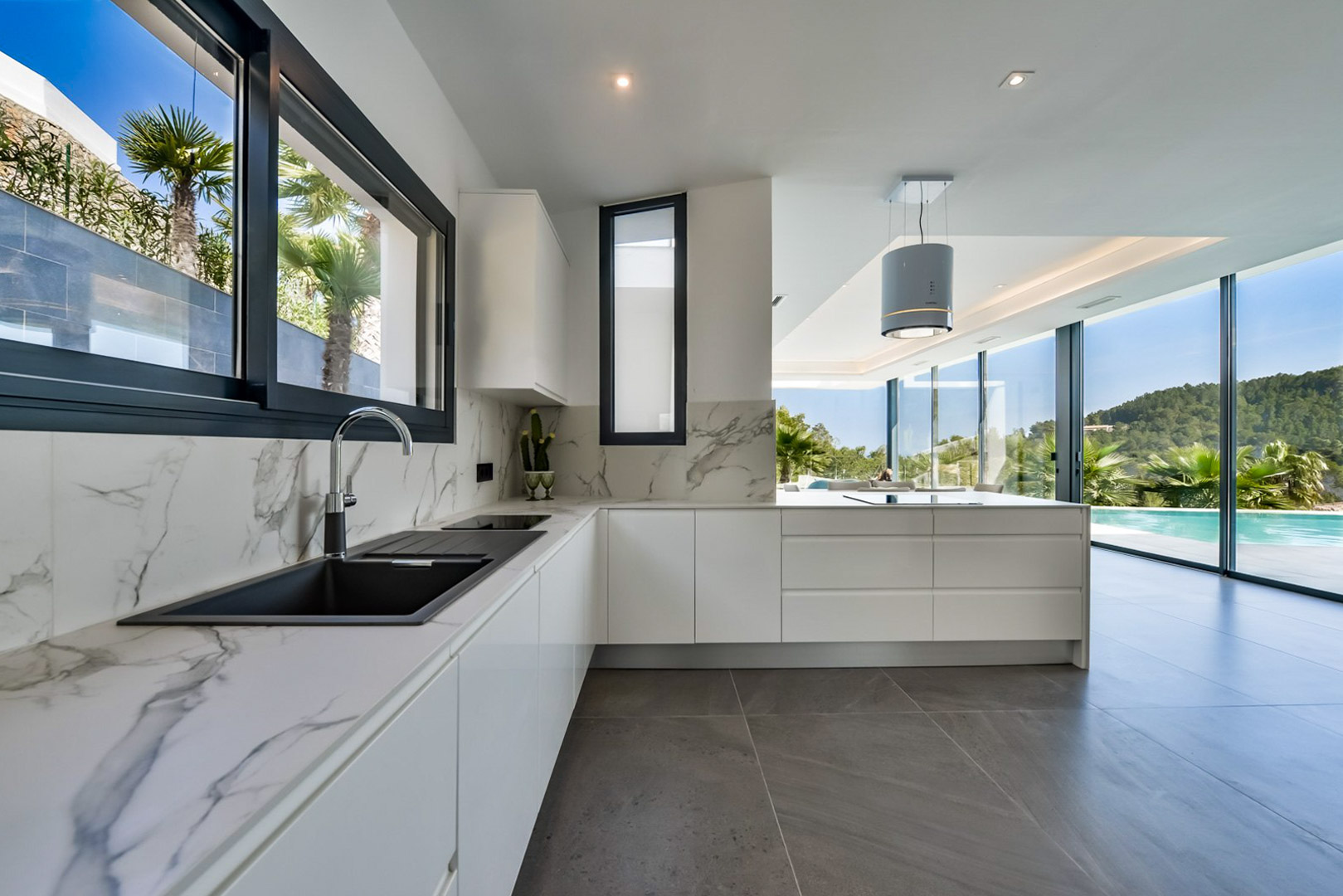 Cocina moderna con encimeras de mármol, fregadero negro y campana cilíndrica. Sus amplios ventanales conectan visualmente el interior con la piscina y el entorno natural.