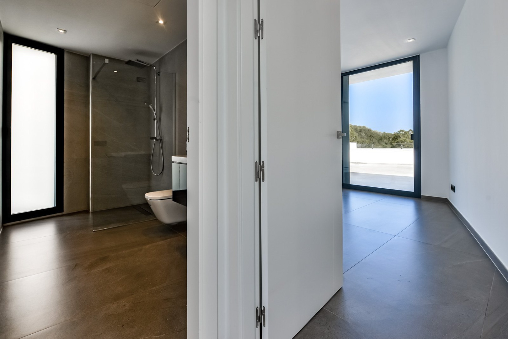 Dormitorio principal con baño en suite, ducha italiana, suelos de gres porcelánico y acceso directo a la terraza mediante amplios ventanales de suelo a techo.