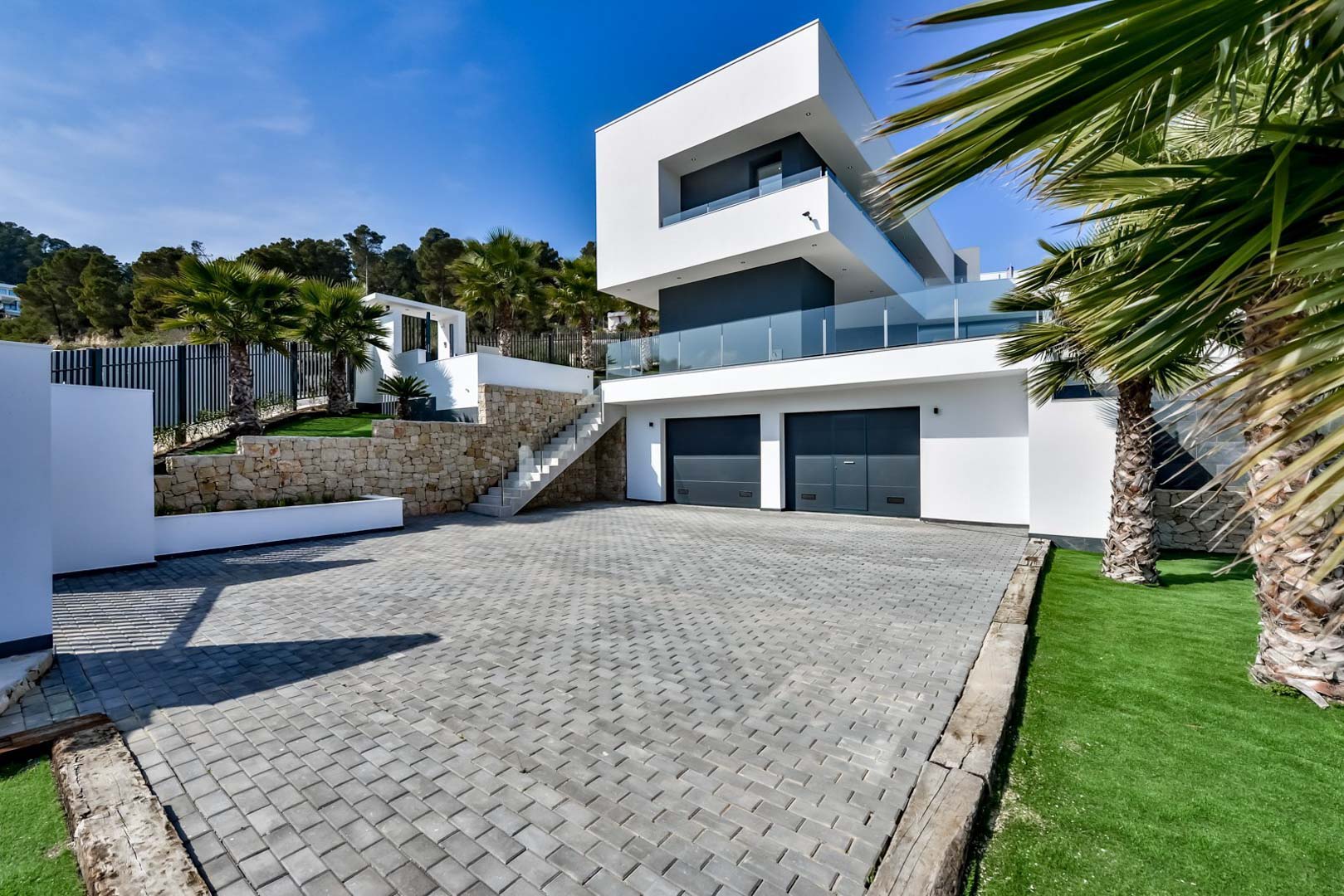 Fachada de diseño contemporáneo con garaje doble, muros de piedra natural y acceso pavimentado rodeado de palmeras y zonas verdes.