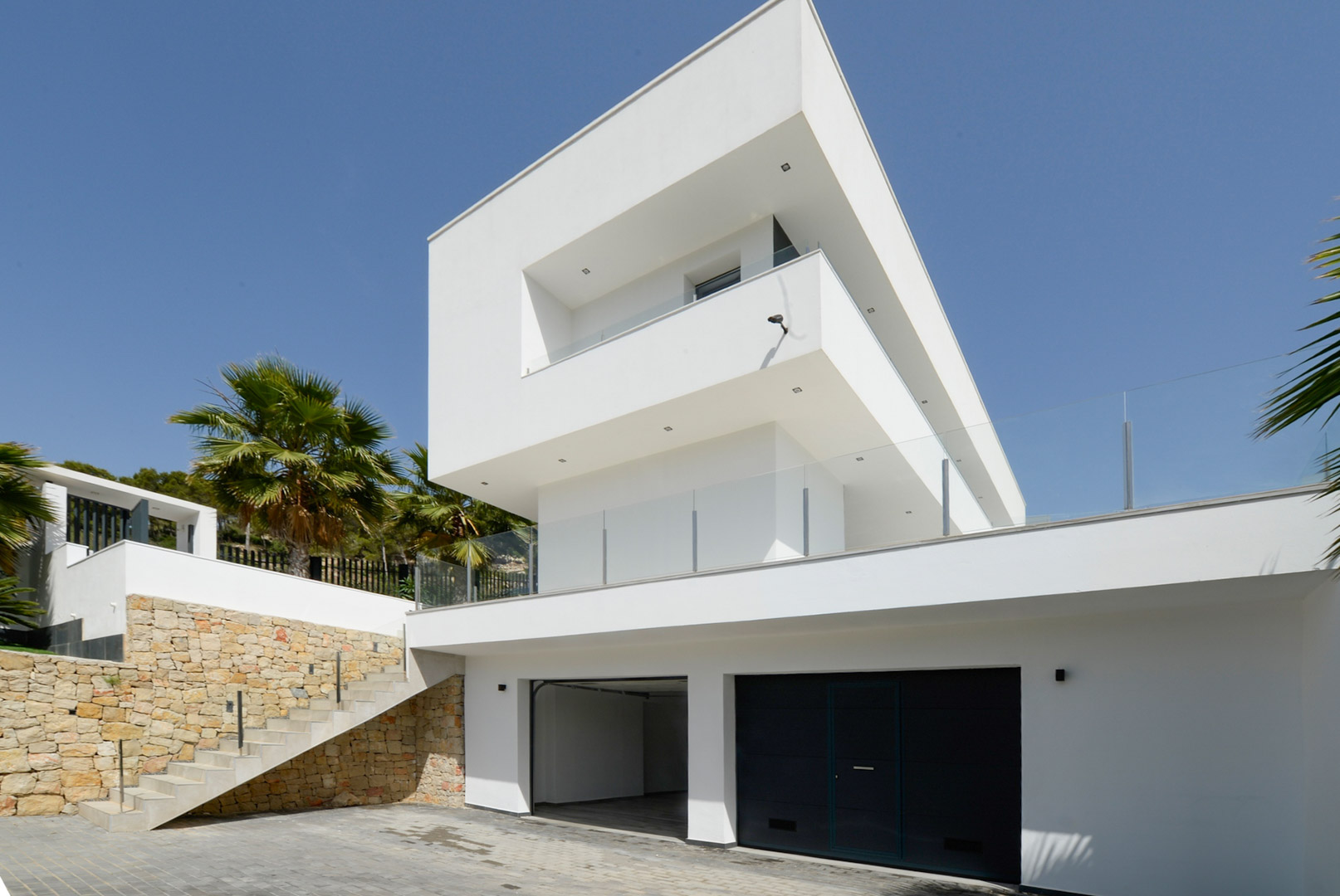 Fachada exterior de arquitectura moderna con volúmenes blancos, garaje doble, muros de piedra natural y escaleras de acceso con barandillas de cristal.