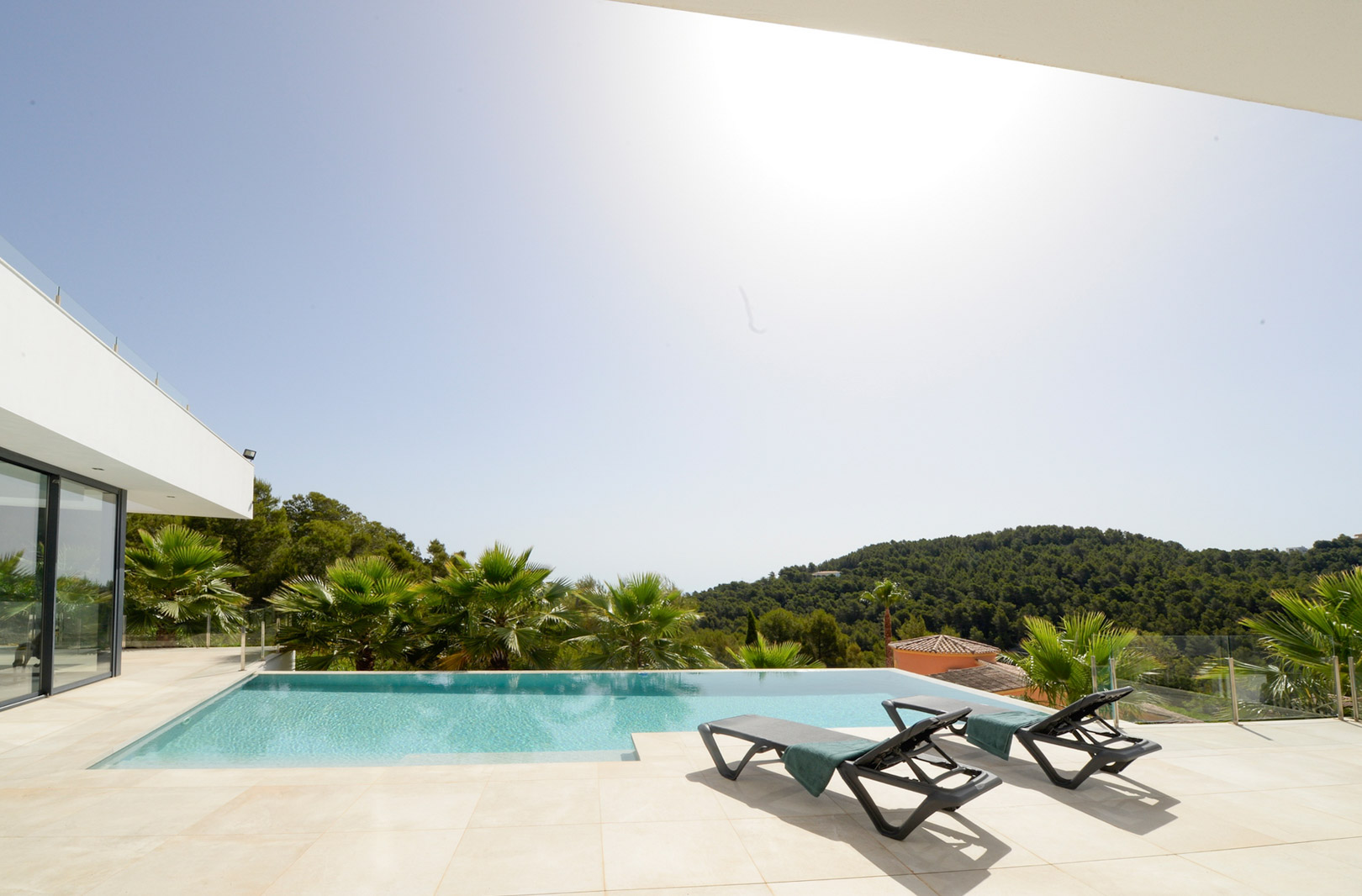 Terraza con piscina infinity, tumbonas y vistas despejadas a las colinas verdes de Jávea, rodeada de palmeras y arquitectura moderna.