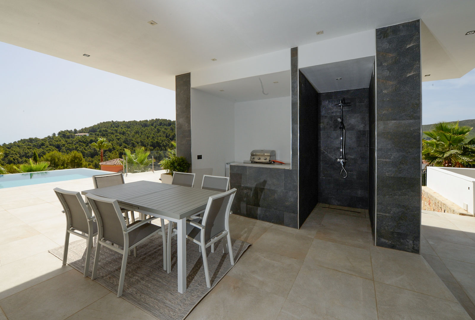 Terraza cubierta con zona de comedor exterior, cocina de verano con barbacoa y ducha exterior revestida en piedra oscura junto a la piscina.