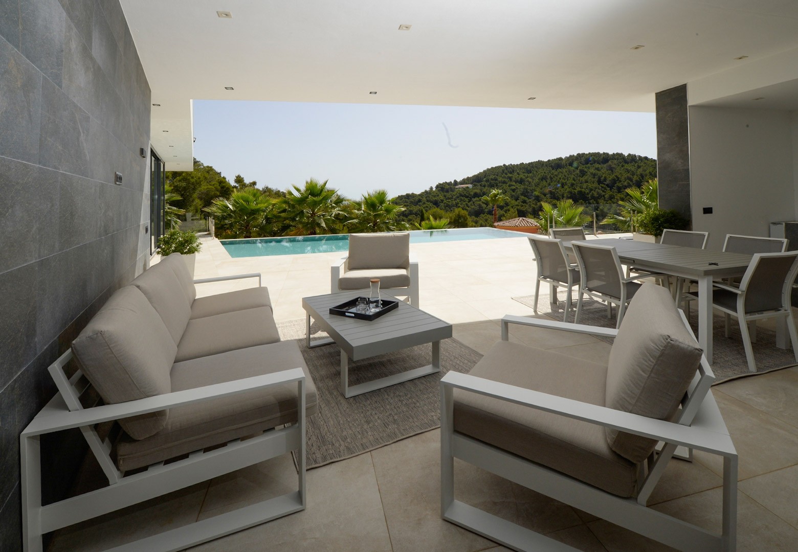 Terraza cubierta con zona de estar y comedor exterior, pavimentos de gres porcelánico y vistas a la piscina infinity y colinas circundantes.