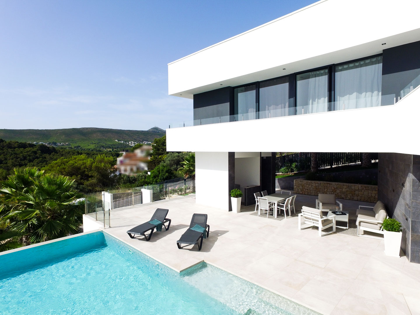 Terraza principal con piscina infinity, zona de estar exterior y comedor bajo el voladizo de la planta superior, con vistas a las colinas de Jávea.