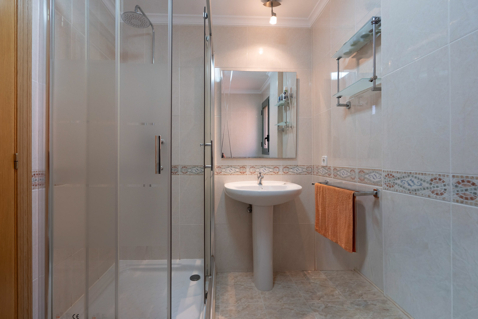 Baño funcional con ducha de obra acristalada, lavabo de pedestal y paredes revestidas con azulejos cerámicos y cenefa decorativa clásica.