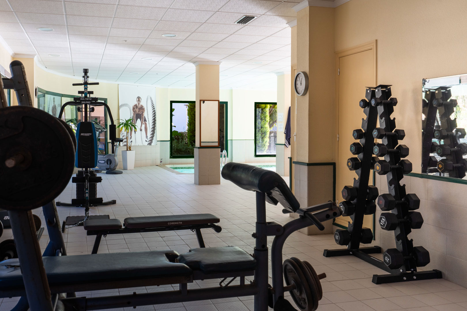 Gimnasio comunitario equipado con máquinas de musculación, pesas y bancos, situado junto a la piscina cubierta con vistas al jardín exterior.