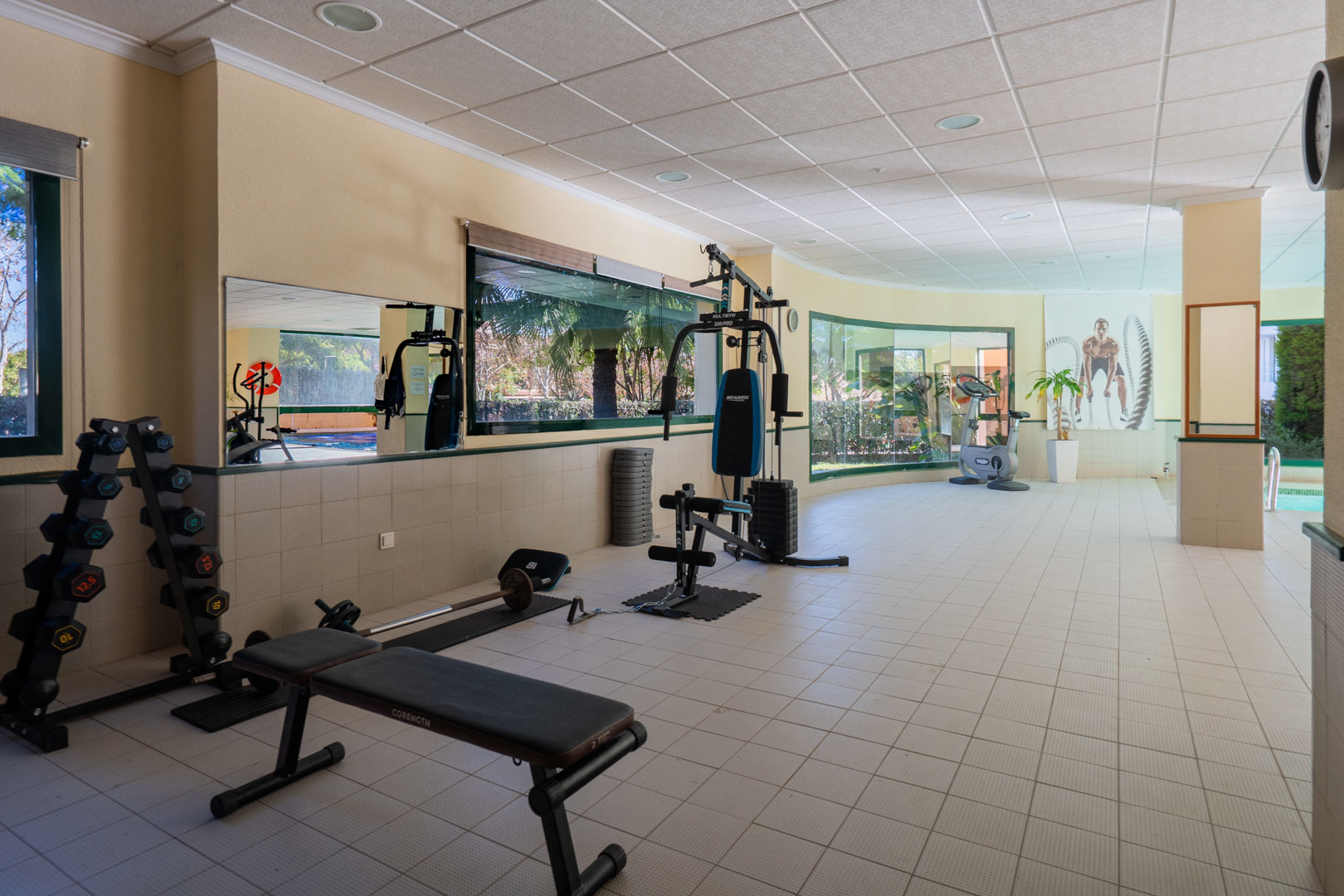 Gimnasio comunitario equipado con máquinas de musculación, pesas y bicicleta estática, con amplios ventanales que ofrecen luz natural y vistas a las zonas verdes.