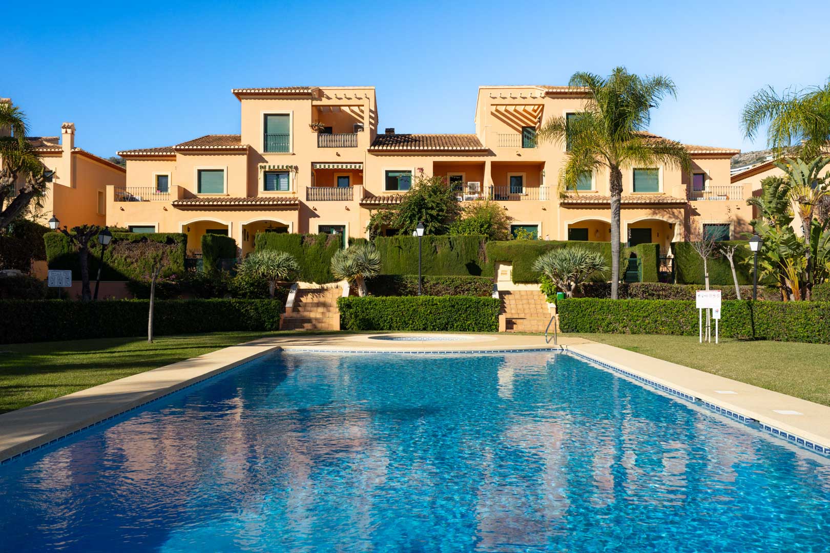Piscina comunitaria rodeada de jardines mediterráneos y césped, con la fachada de estilo tradicional del complejo Floridamar al fondo bajo un cielo despejado.