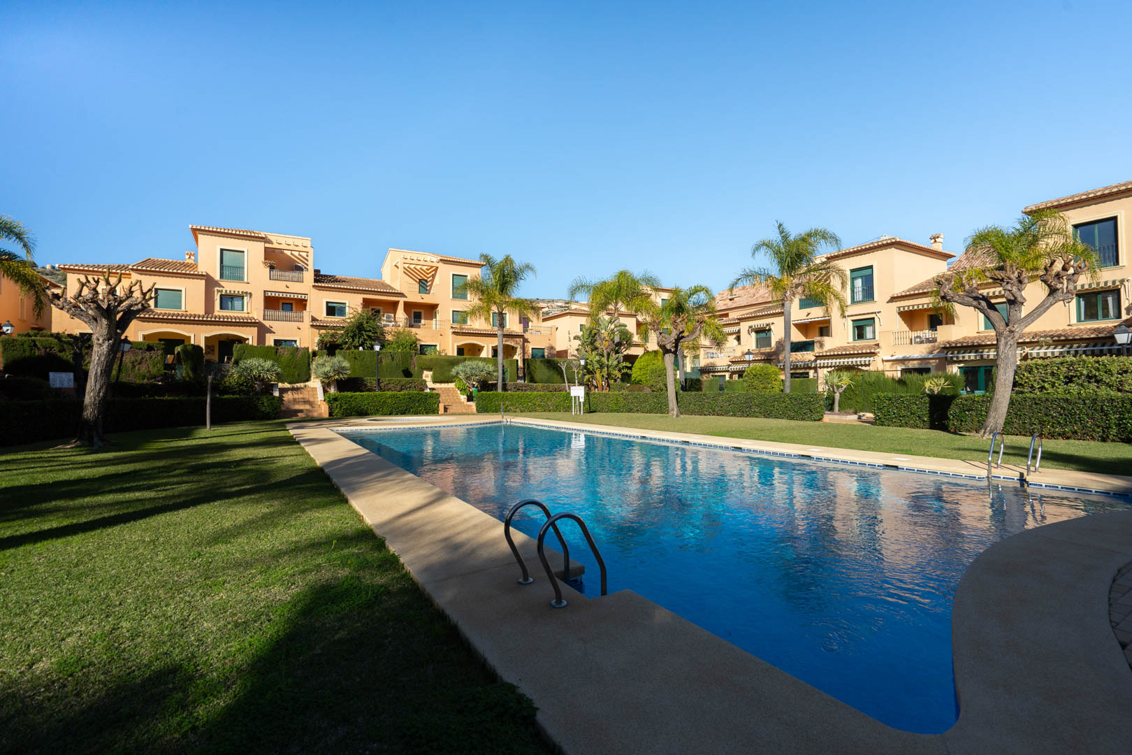 Piscina exterior comunitaria rodeada de amplias zonas de césped, palmeras y edificios de estilo mediterráneo bajo un cielo despejado en Floridamar.
