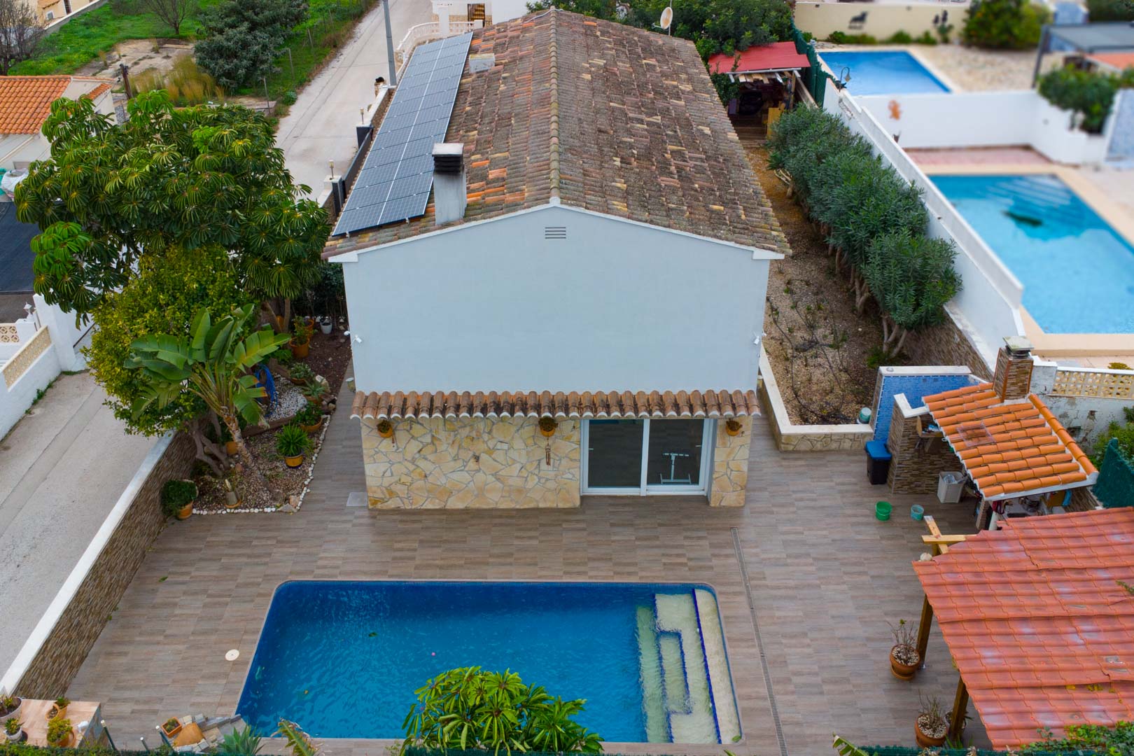 Vista aérea de la villa con paneles solares en el tejado, piscina privada y terraza pavimentada con zona de barbacoa cubierta.