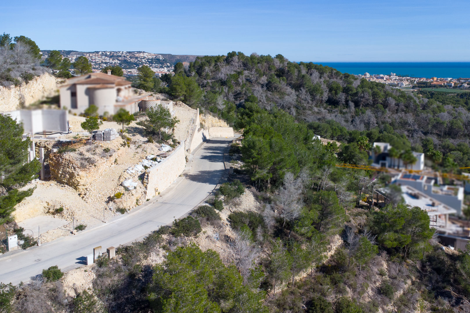 Vista aérea de la calle de acceso asfaltada en Tosalet 5, rodeada de pinos y muros de contención de piedra, con el mar Mediterráneo y la costa de Jávea al fondo.