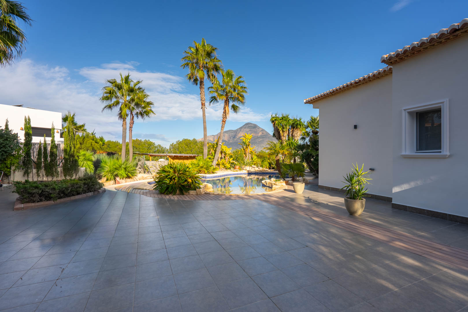 Amplia terraza exterior con pavimento cerámico gris, piscina de diseño orgánico rodeada de palmeras y vistas despejadas al macizo del Montgó bajo un cielo despejado.