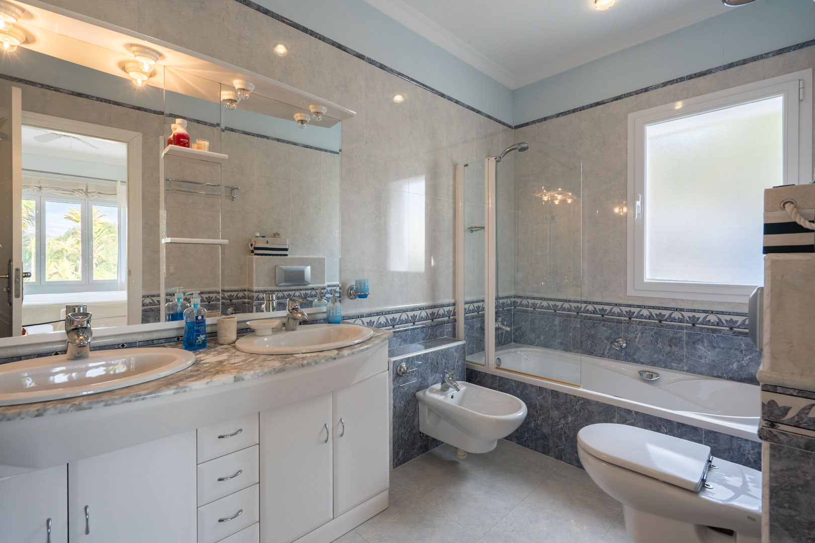 Baño completo con lavabo de doble seno sobre encimera de mármol, bañera con mampara de cristal y azulejos en tonos azules y grises con cenefas decorativas.