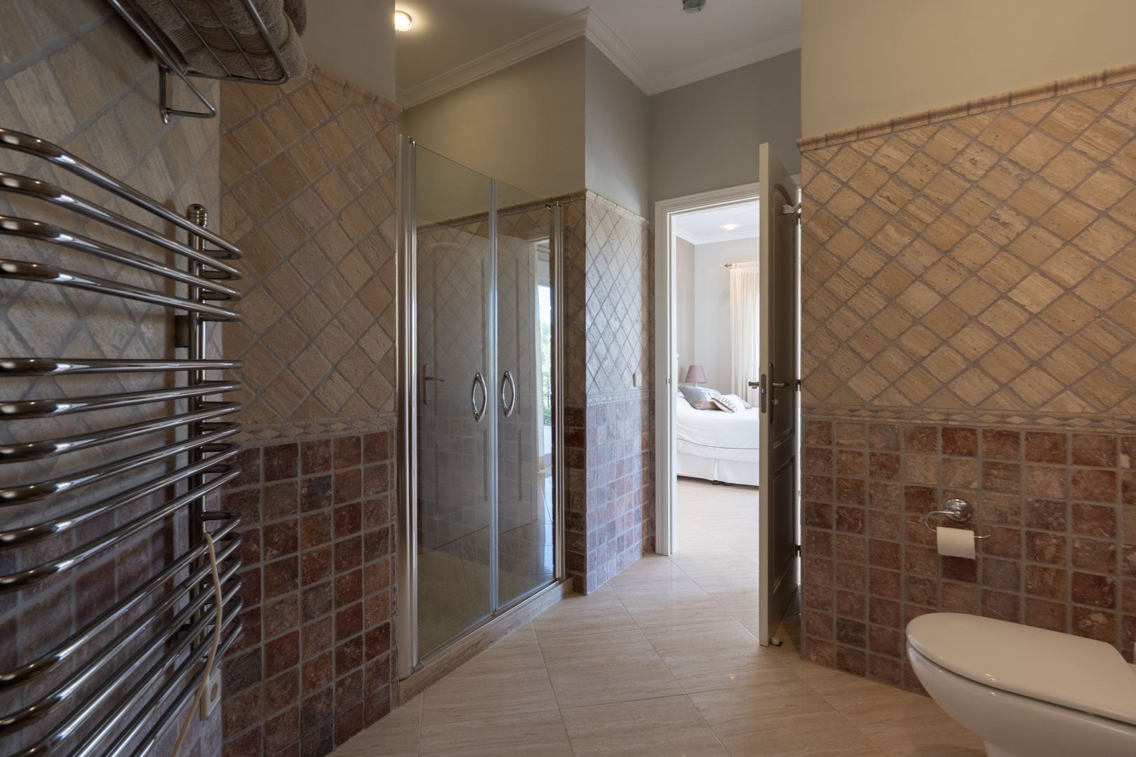 Baño en suite con ducha de cristal, toallero calefactado cromado y azulejos en tonos tierra, conectado directamente con un dormitorio luminoso.