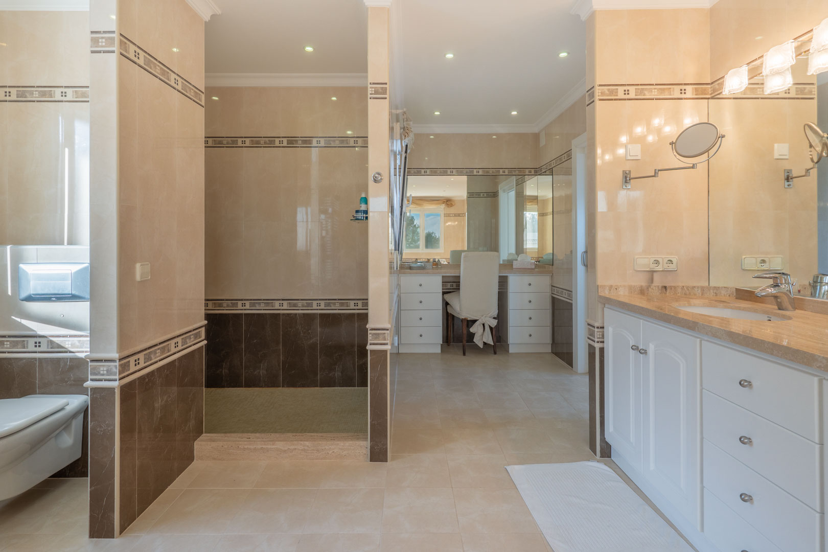Baño principal con azulejos en dos tonos, ducha de obra, zona de tocador con espejo y lavabo encastrado en encimera de mármol con mobiliario blanco.