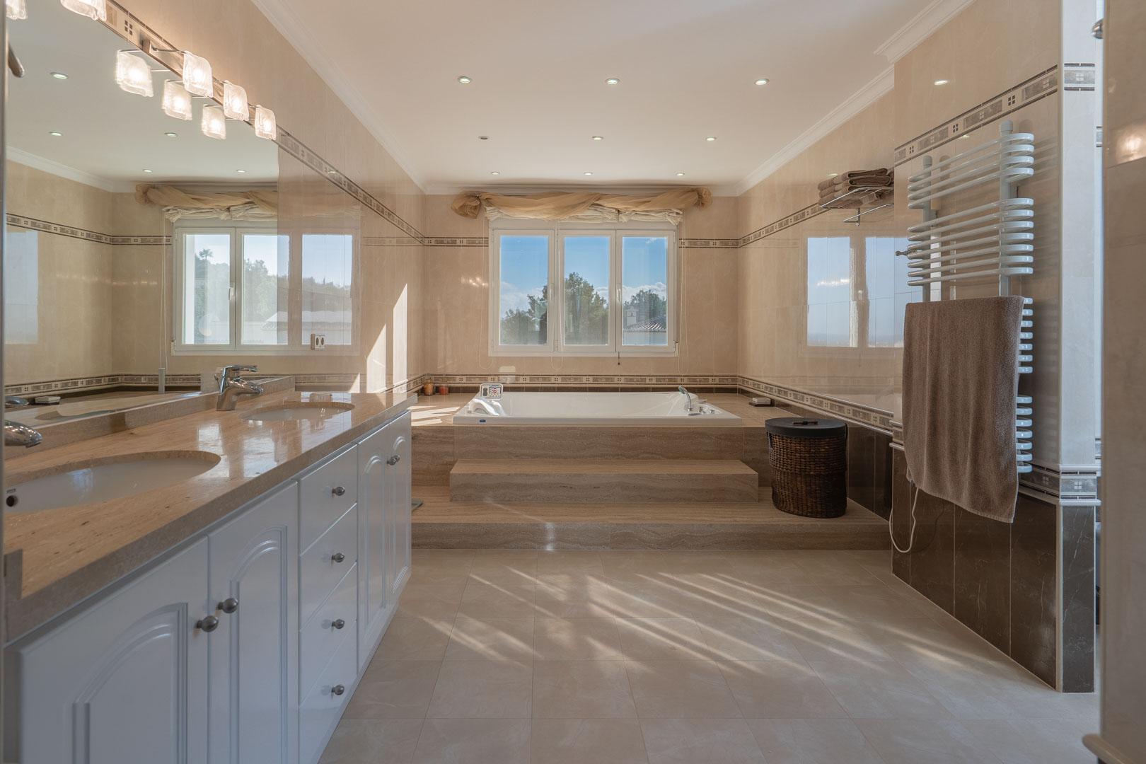 Baño principal con encimera de mármol doble, bañera de hidromasaje elevada sobre peldaños de piedra y acabados en tonos crema con luz natural.