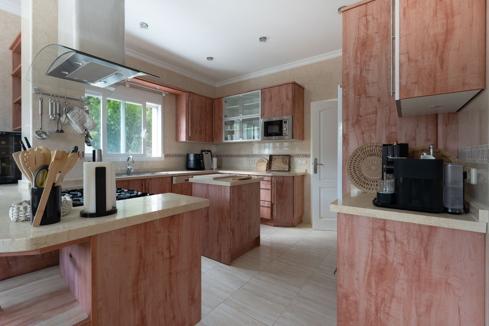 Cocina equipada con mobiliario de madera rojiza, encimeras de piedra clara, isla central y campana de cristal sobre la zona de cocción a gas.