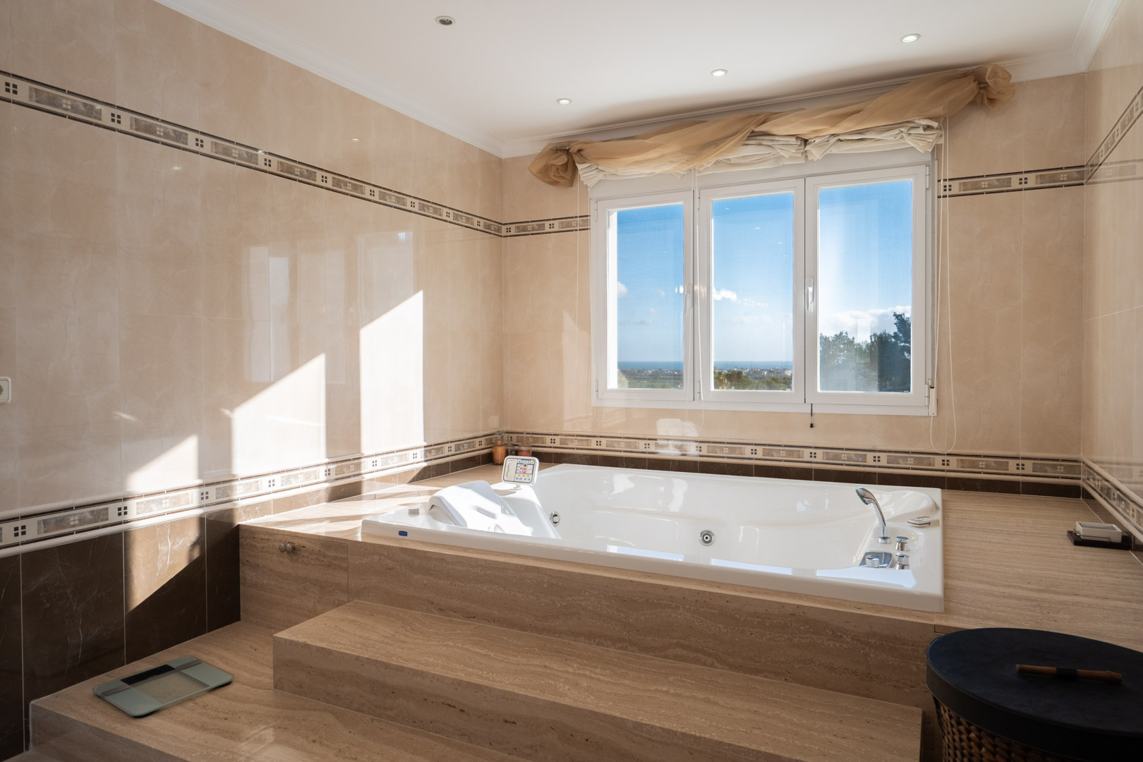Cuarto de baño con bañera de hidromasaje encastrada en mármol, ventanal con vistas al mar y revestimientos cerámicos en tonos crema con cenefas decorativas.