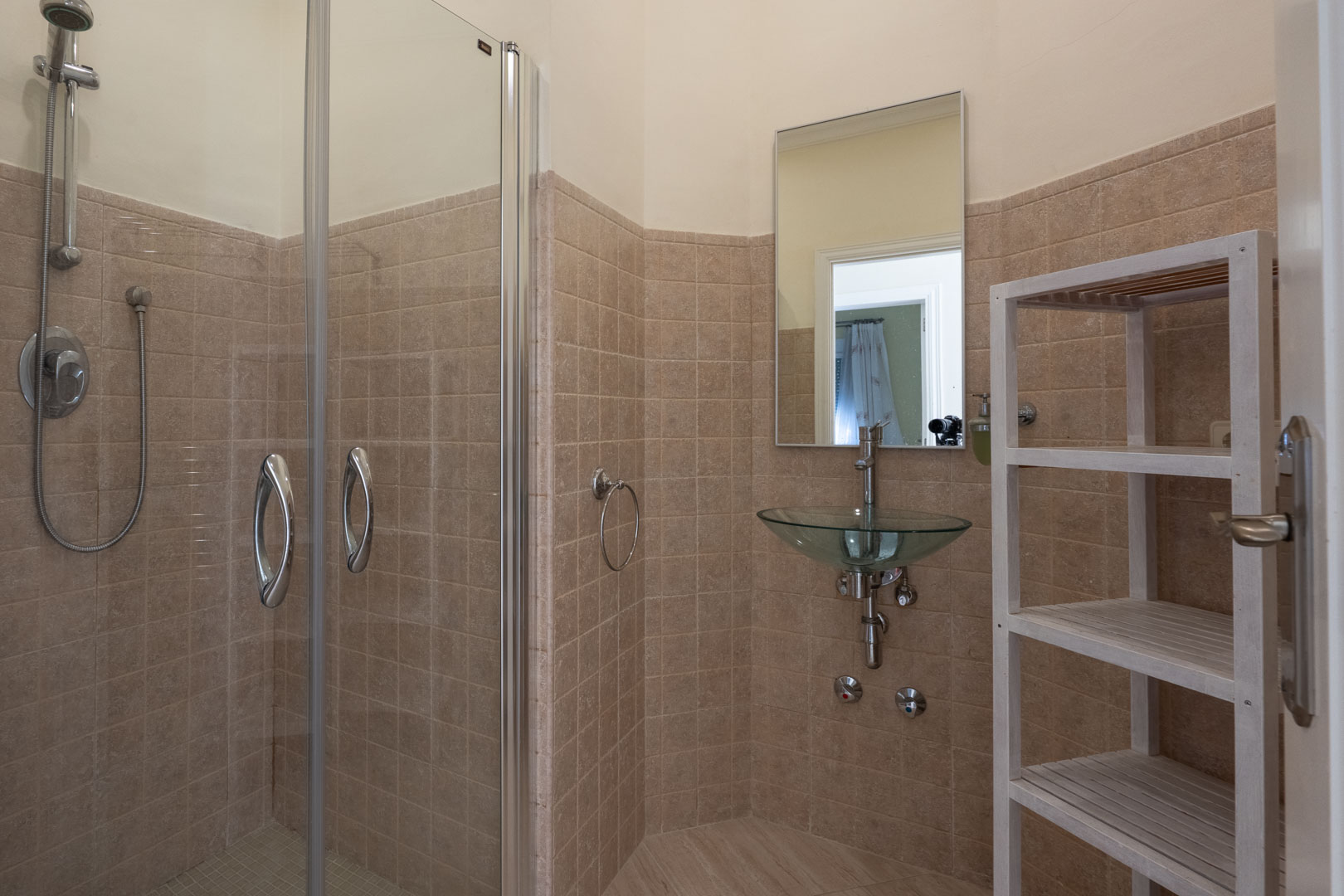 Cuarto de baño con ducha de obra acristalada, lavabo de cristal de diseño suspendido, paredes revestidas en gres beige y estantería auxiliar de madera blanca.