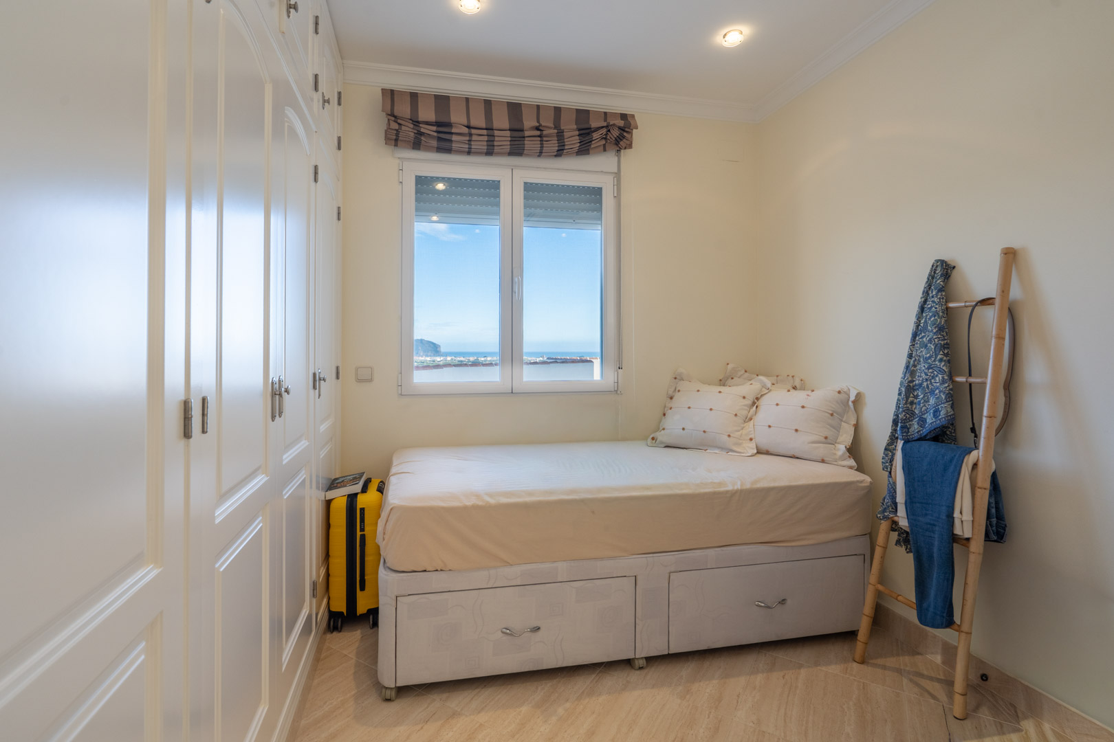 Dormitorio con armarios empotrados lacados en blanco, cama con almacenaje inferior y ventana con vistas directas al mar Mediterráneo.