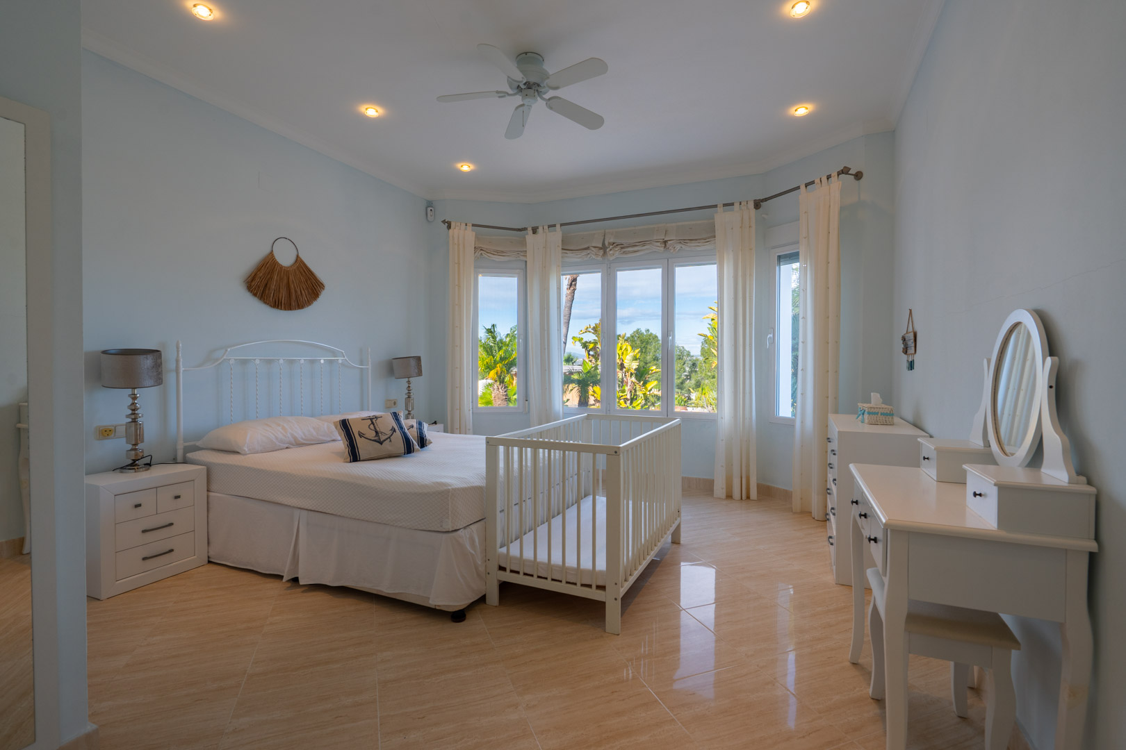 Dormitorio con suelos de gres brillante, ventanal con vistas al jardín, mobiliario blanco, tocador y ventilador de techo en un ambiente sereno.