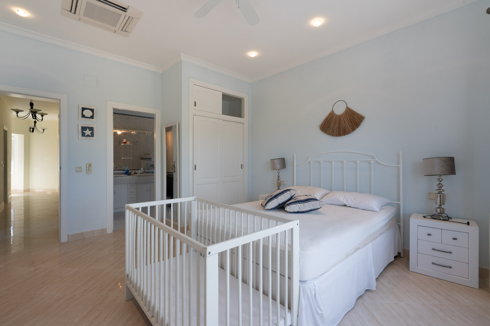Dormitorio luminoso con paredes en azul pálido, cama doble con cabecero de forja blanca, cuna integrada, armario empotrado y acceso directo a un baño en suite.