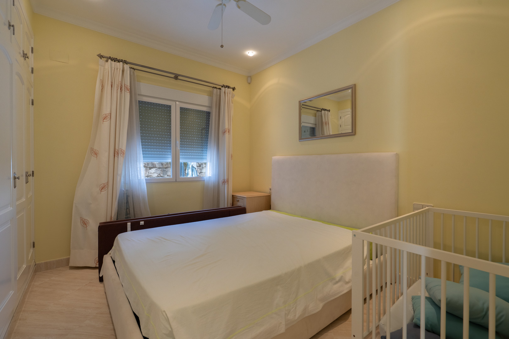 Dormitorio luminoso con paredes en tono crema, armarios empotrados blancos, cama doble con cabecero tapizado y cuna. Incluye ventilador de techo y ventana con persiana.