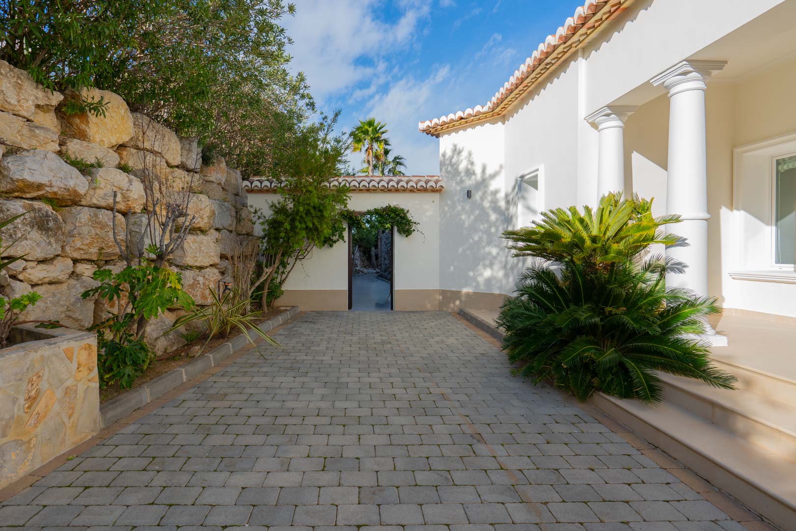 Entrada exterior con suelo de adoquines grises, muro de piedra natural y fachada blanca con columnas clásicas junto a vegetación mediterránea.