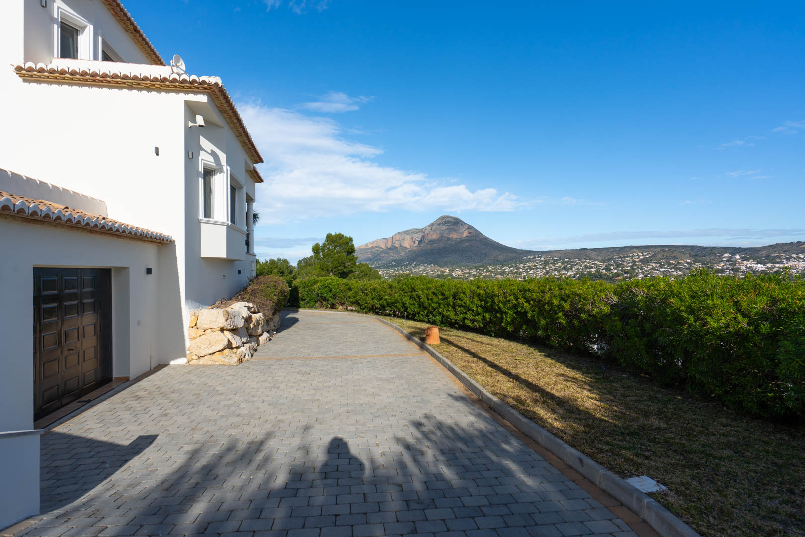 Entrada pavimentada con acceso al garaje, flanqueada por muros de piedra natural y setos, con vistas despejadas al macizo del Montgó.