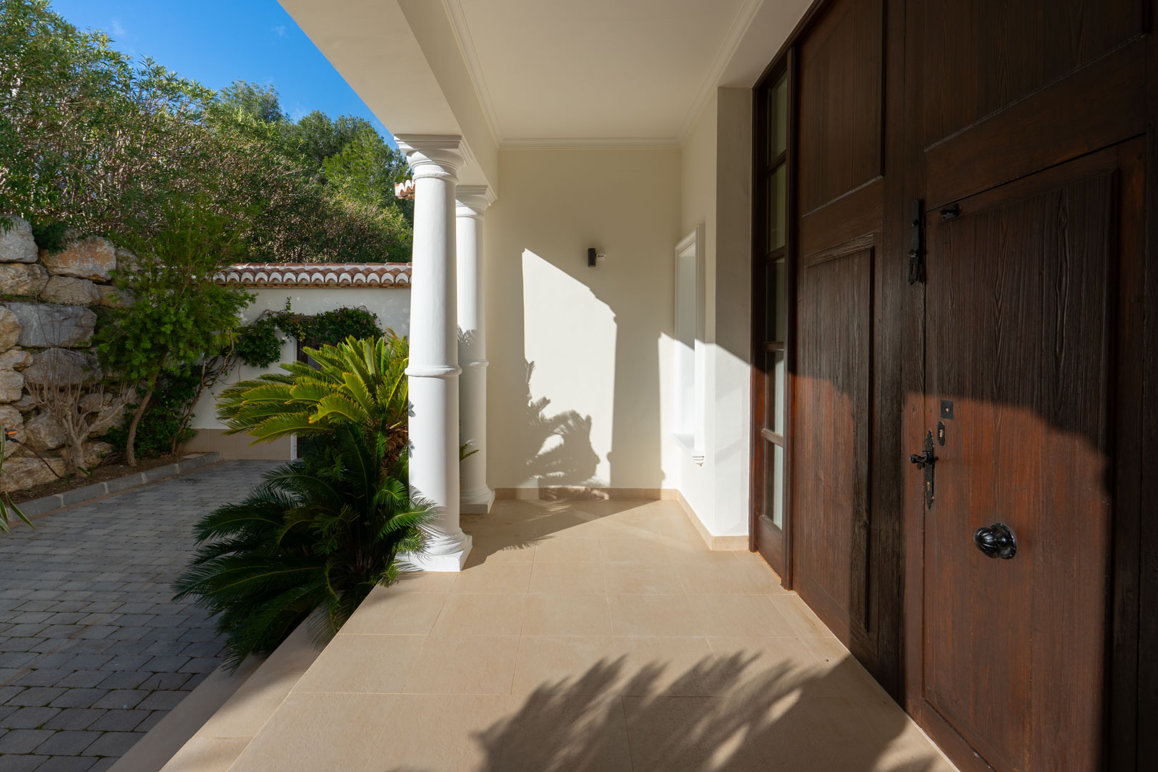 Entrada principal con puerta de madera maciza, columnas blancas de estilo clásico y porche de piedra natural rodeado de vegetación mediterránea.