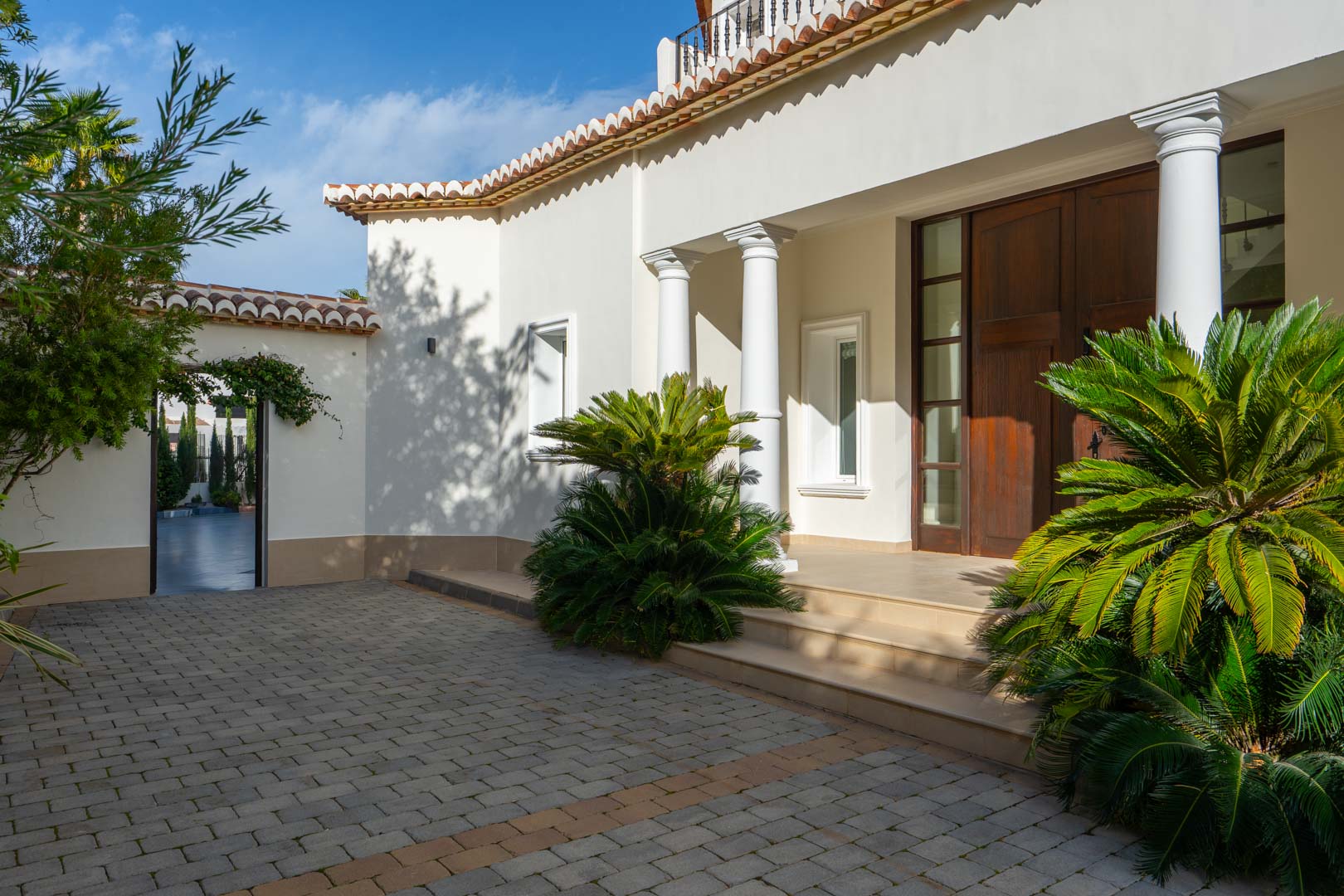 Entrada principal con puerta de madera noble, columnas blancas y zona de acceso pavimentada rodeada de vegetación mediterránea.