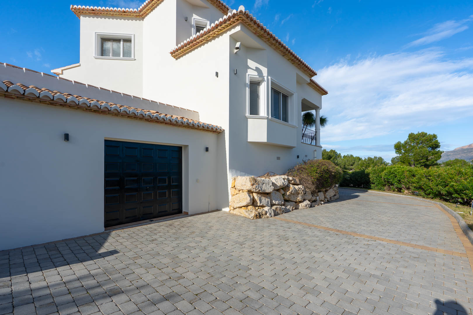 Entrada principal pavimentada con acceso a garaje privado, destacando la fachada blanca de estilo mediterráneo con muros de contención en piedra natural.